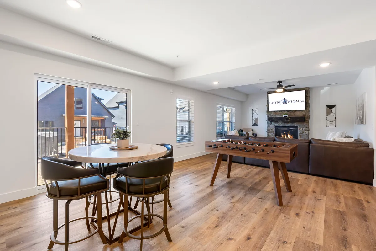 A spacious lower-level living area featuring a cozy stone fireplace, a foosball table ready for lively competitions, a modern dining table for games or snacks, and a comfortable sofa arrangement. The ambiance is playful yet inviting, perfectly blending relaxation with entertainment.