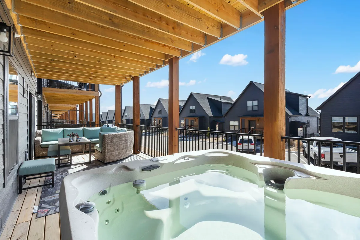 An exterior patio space with a hot tub, ample outdoor seating, wooden beams overhead, and nearby houses in the background. The area is set up for socializing and relaxation, evoking a sense of community living and shared enjoyment of the outdoors.
