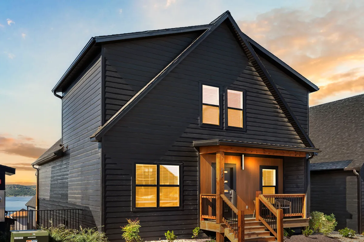 An inviting exterior view of the property with dark wooden siding, large windows, and a porch set against a glowing sunset sky. The porch is ideal for relaxing and soaking in the sights and sounds of nature.
