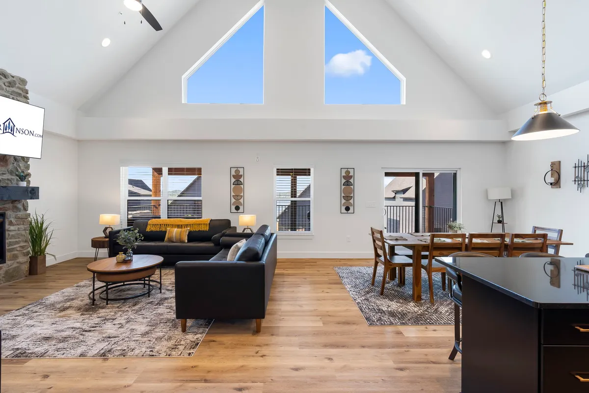 The hero image showcases a spacious, sun-drenched living area featuring a plush sofa, a stylish dining table perfect for group meals, a cozy fireplace, and expansive windows that offer sweeping views of Table Rock Lake. The room feels warm and welcoming, ideal for family gatherings or relaxing with friends.