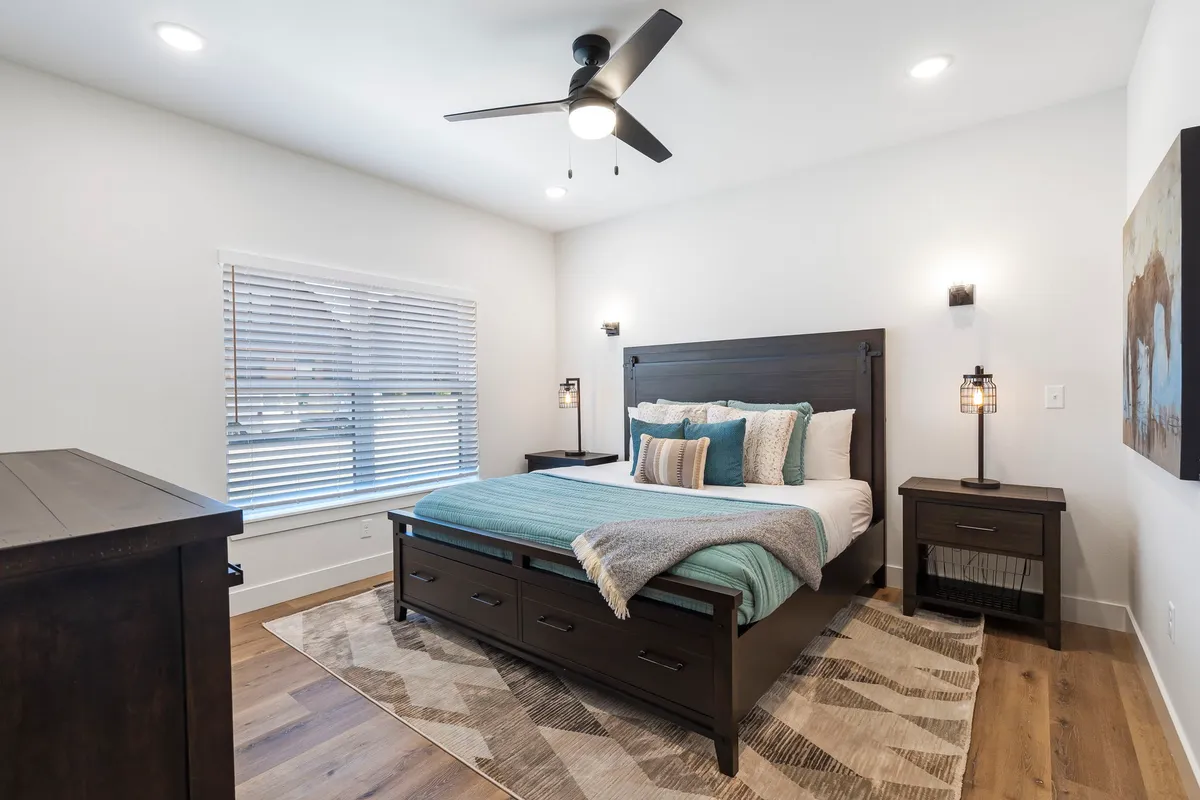 A cozy bedroom featuring a bed with decorative pillows, nightstands with lamps on either side, a window with blinds letting in natural light, and a soft rug on the floor. The neutral color palette and simple decor create an inviting, restful atmosphere.