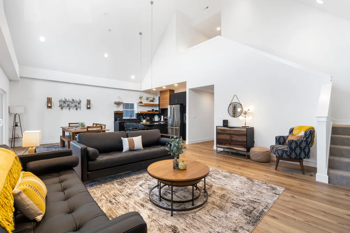 A wide view of the main living area, seamlessly connecting the sofa, coffee table, dining area, and kitchen, with a staircase visible. The space is bathed in warm natural light, conveying a sense of openness and comfort ideal for gatherings.
