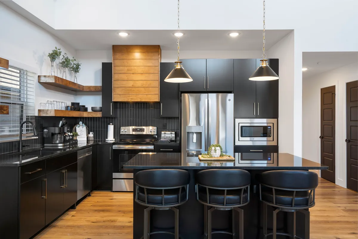 A modern kitchen featuring a spacious island, sleek black cabinetry, wood accents, and up-to-date appliances. The space is set up for both cooking and socializing, with plenty of room for multiple people to gather.